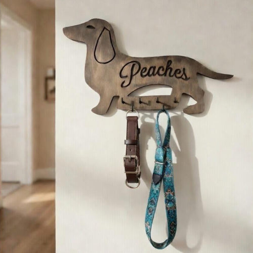 Wooden dog-shaped leash holder with 'Peaches' engraved, holding a collar and leash against a white wall.